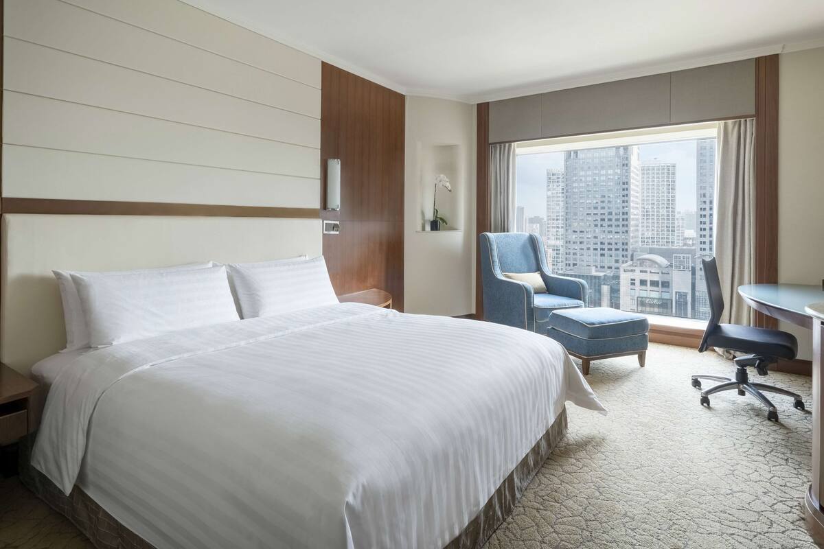 A hotel room with a large bed, a desk, a chair, and a view of tall buildings.