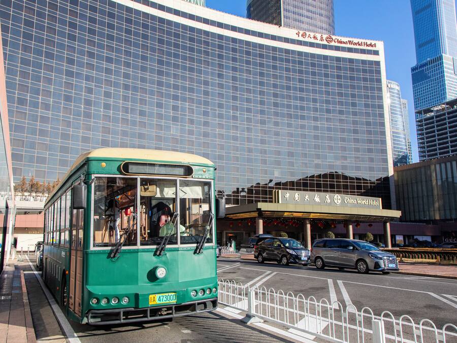 A green bus at a bus stop in front of a modern building with a China World Hotel sign.