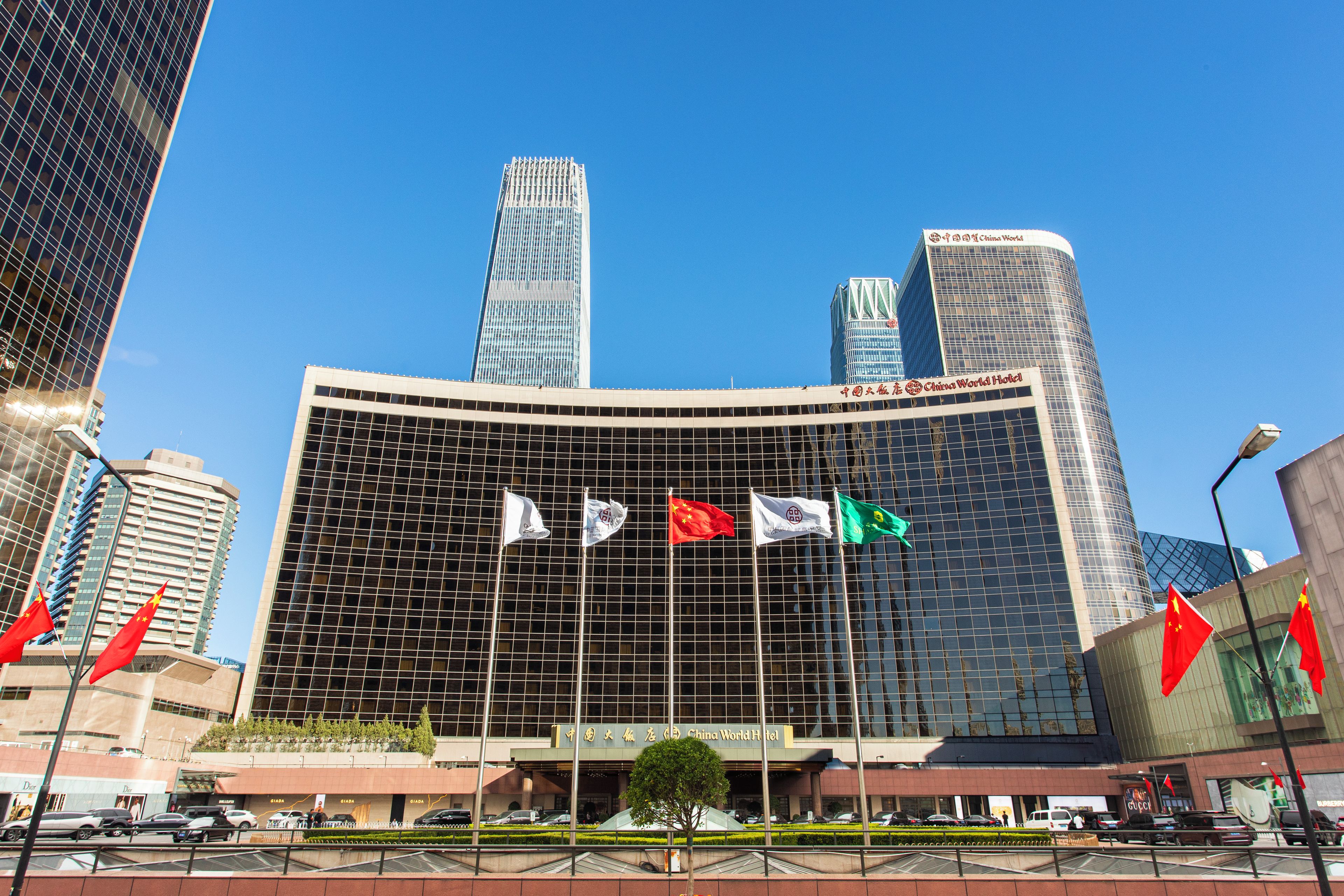 Un moderno edificio de hotel con varias banderas, incluyendo la bandera china, y un cielo azul despejado al fondo.