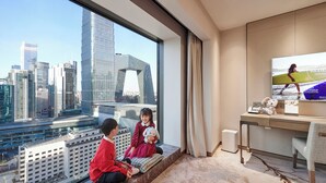 Ein modernes Hotelzimmer mit großem Fenster und Blick auf die Stadt, einem Schreibtisch mit Computer sowie einer gemütlichen Sitzecke für zwei Personen.