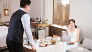 Ein Hotelzimmer mit einem Bett, einem Schreibtisch mit Laptop, einem Esstisch mit Frühstück und einem großen Fenster mit Blick auf die Stadt.