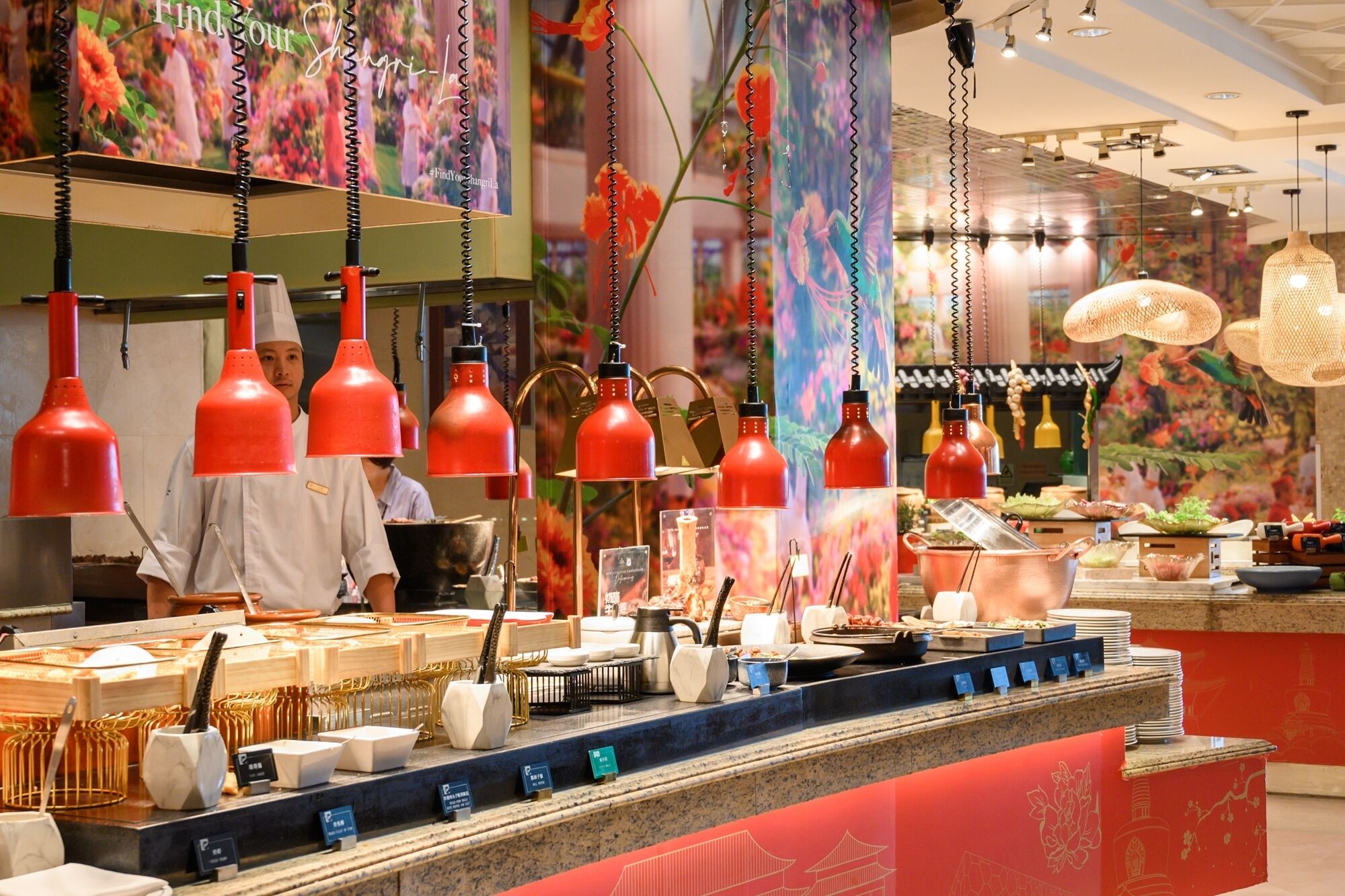 A restaurant interior with red pendant lights, a counter with various dishes and utensils, and a decorative wall with floral patterns.
