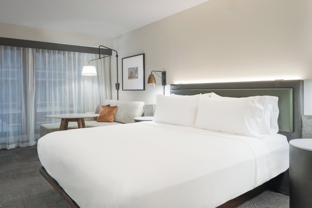 A hotel room with a large bed, a bedside table, a chair, a lamp, and a window with curtains.