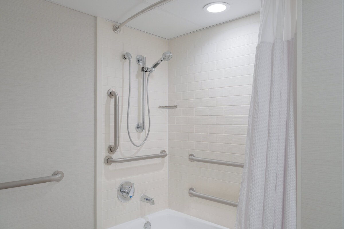 A bathroom with white tiled walls, a white bathtub, and a shower with multiple faucets and a handheld showerhead.