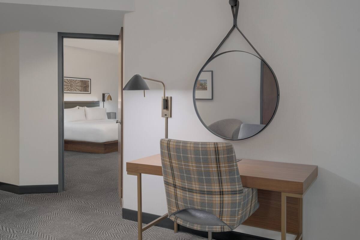 Una habitación de hotel con una cama, un escritorio con silla, un espejo montado en la pared y una lámpara.