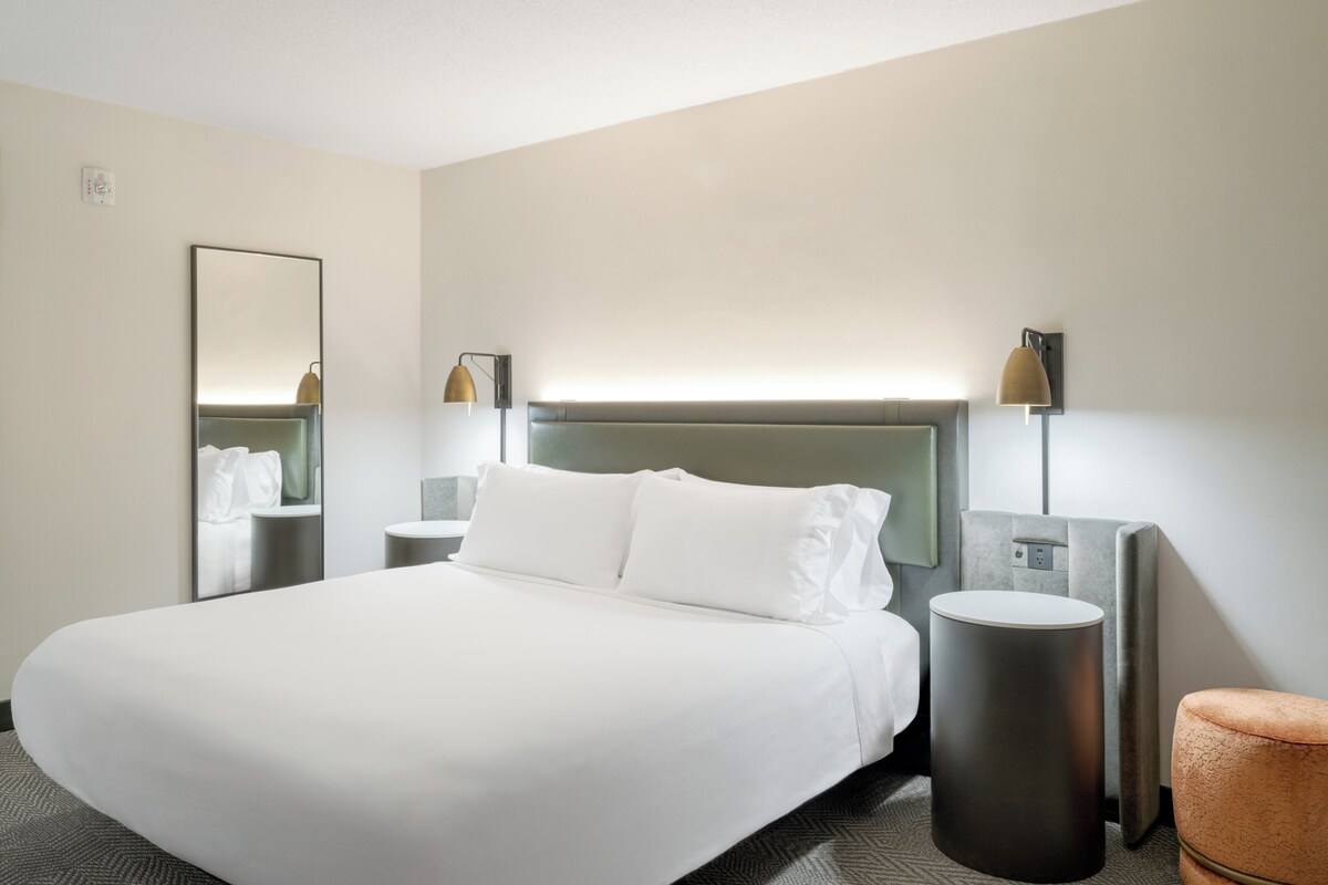 A modern hotel room with a large bed, bedside tables, a round nightstand, a mirror, and a lamp.
