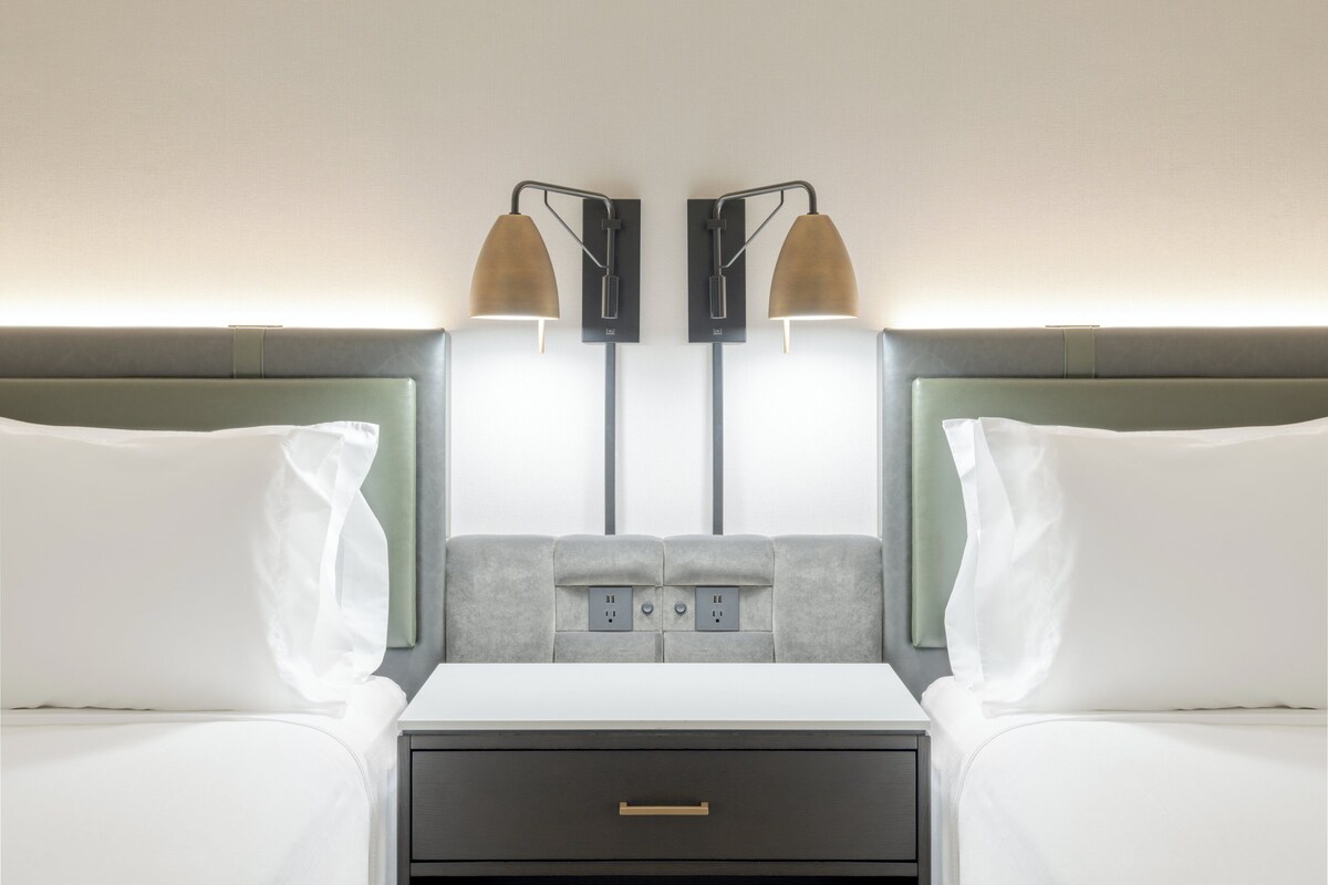 A hotel room with two beds, a nightstand, and a wall-mounted lamp.