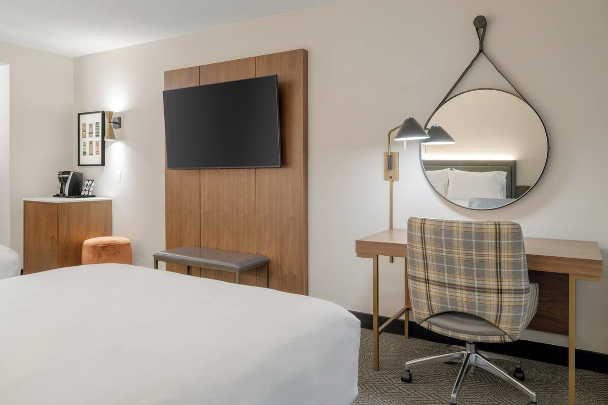 Una habitación de hotel con una cama grande, un escritorio con silla, un televisor montado en la pared, un taburete pequeño y un cuadro enmarcado.
