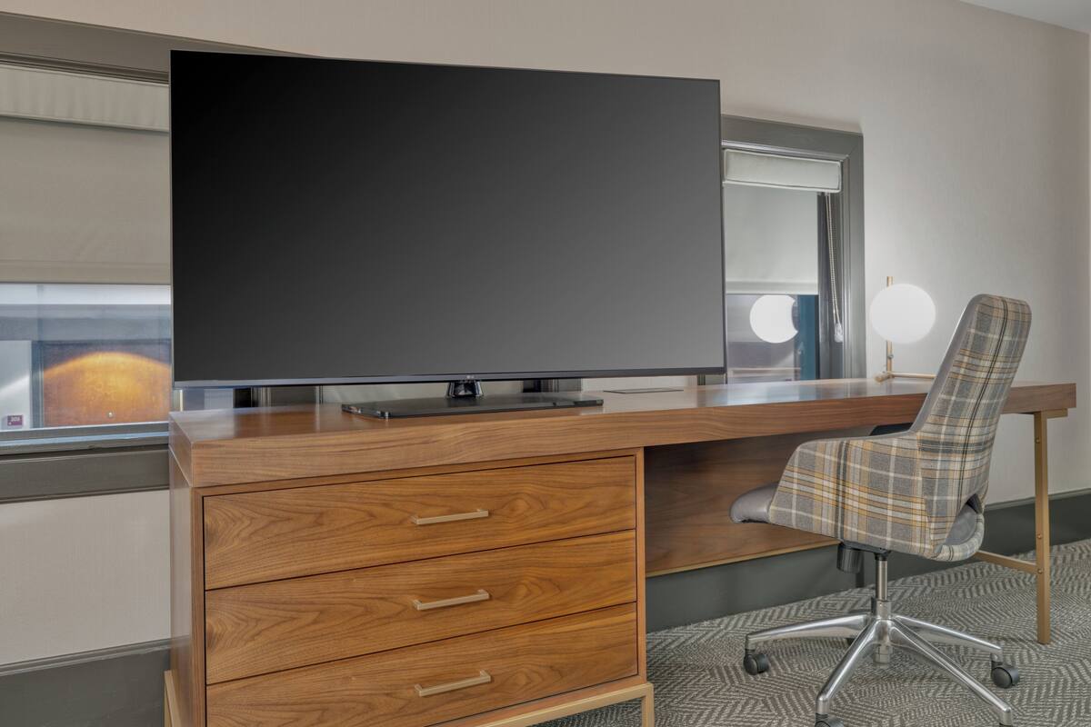Una habitación de hotel moderna con un escritorio de madera, un televisor de pantalla plana grande, un sillón a cuadros y una ventana con persianas.