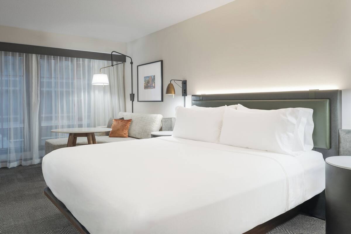 Una habitación de hotel con una cama grande, una mesita de noche, una silla, una lámpara y una ventana con cortinas.
