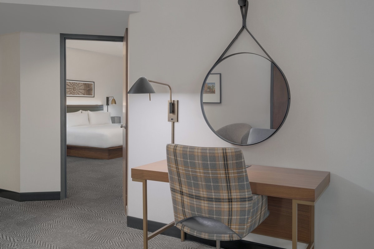 A hotel room with a bed, a desk with a chair, a wall-mounted mirror, and a lamp.