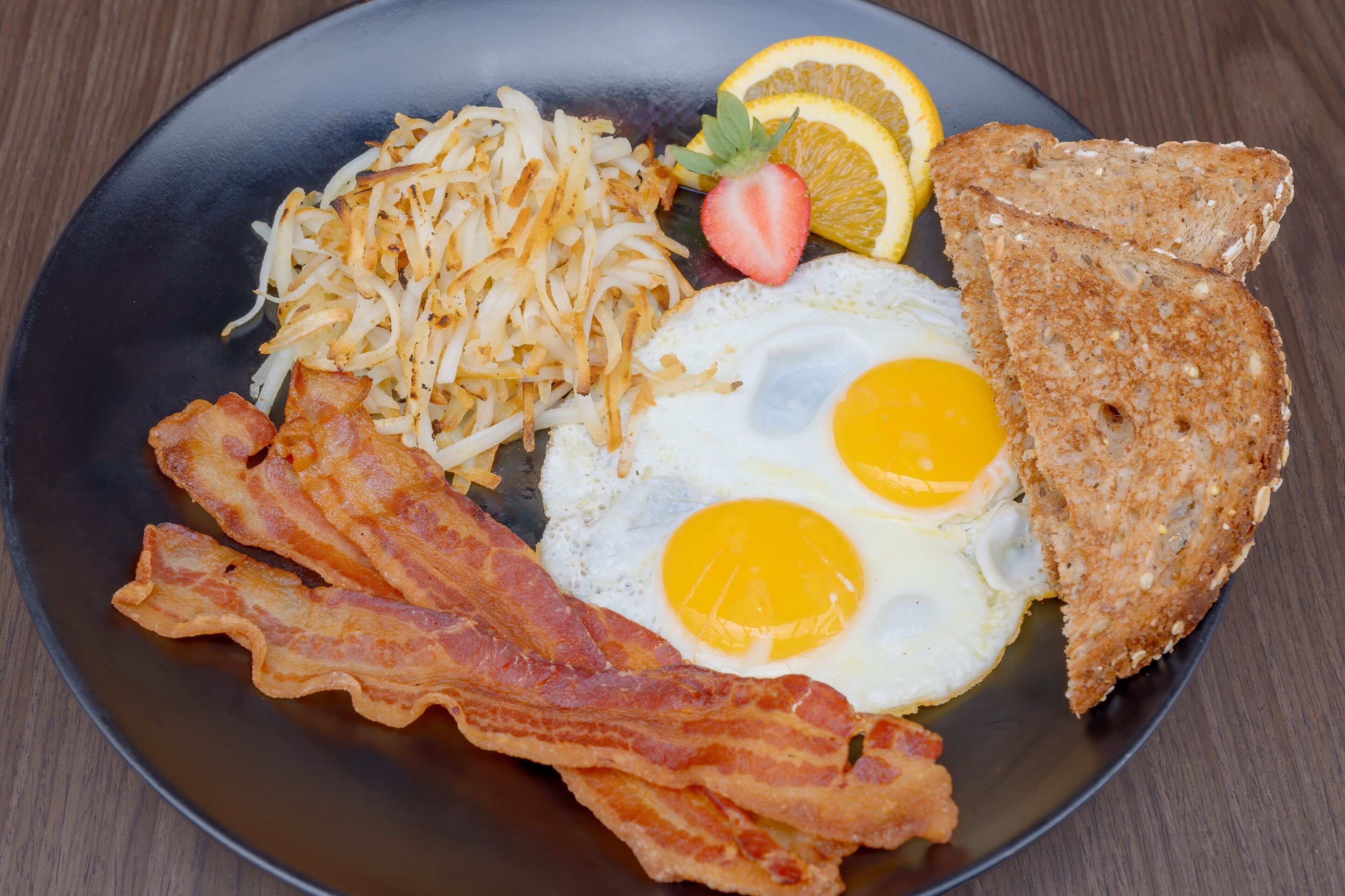 A plate with eggs, bacon, hash browns, toast, a strawberry, and orange slices.