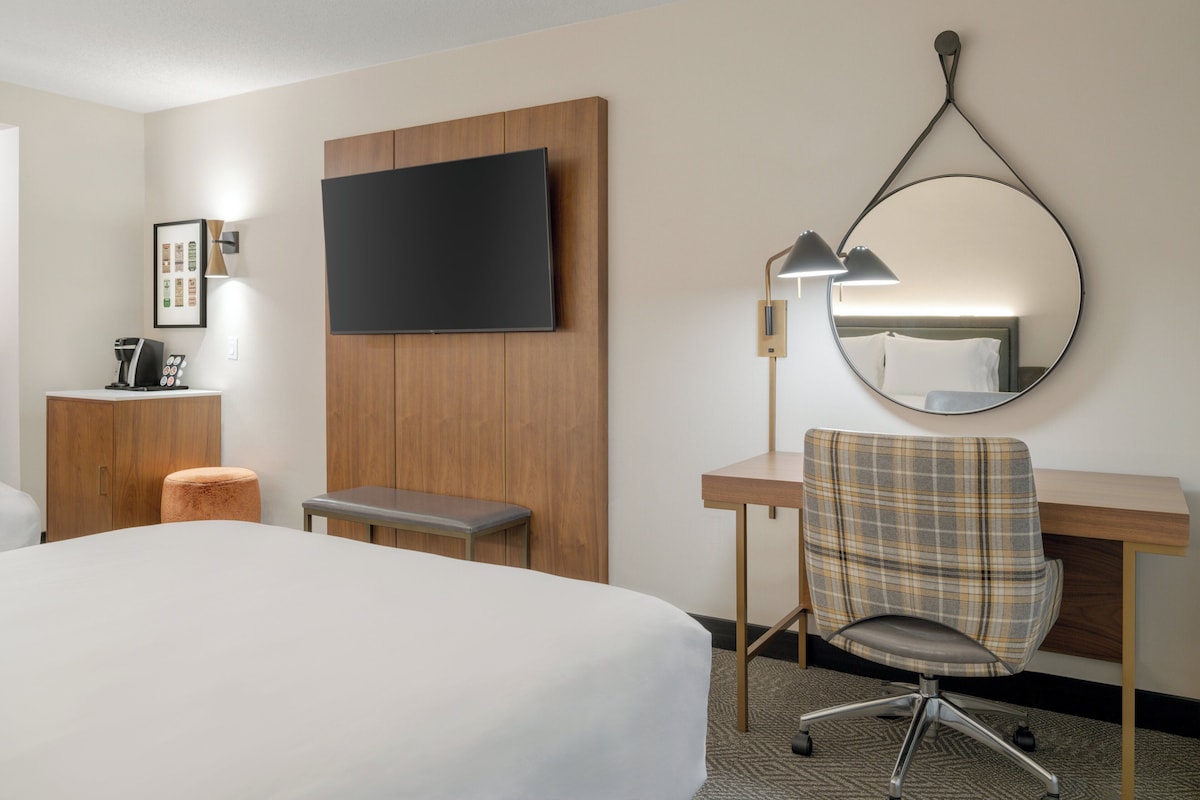 A hotel room with a large bed, a desk with a chair, a television mounted on the wall, a small stool, and a framed picture on the wall.