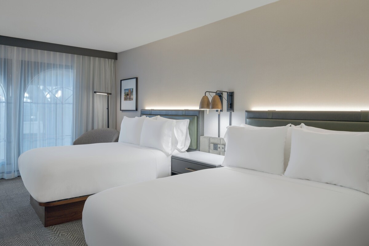 A hotel room with two beds, a desk, a chair, a lamp, and a window with curtains.