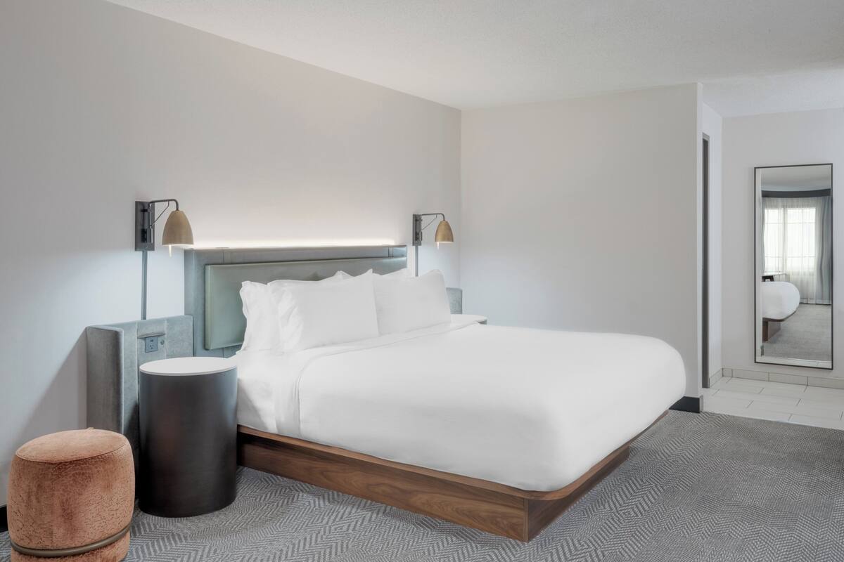 Una habitación de hotel moderna con una cama grande, un reposapiés redondo, una mesita de noche y un espejo en la pared.