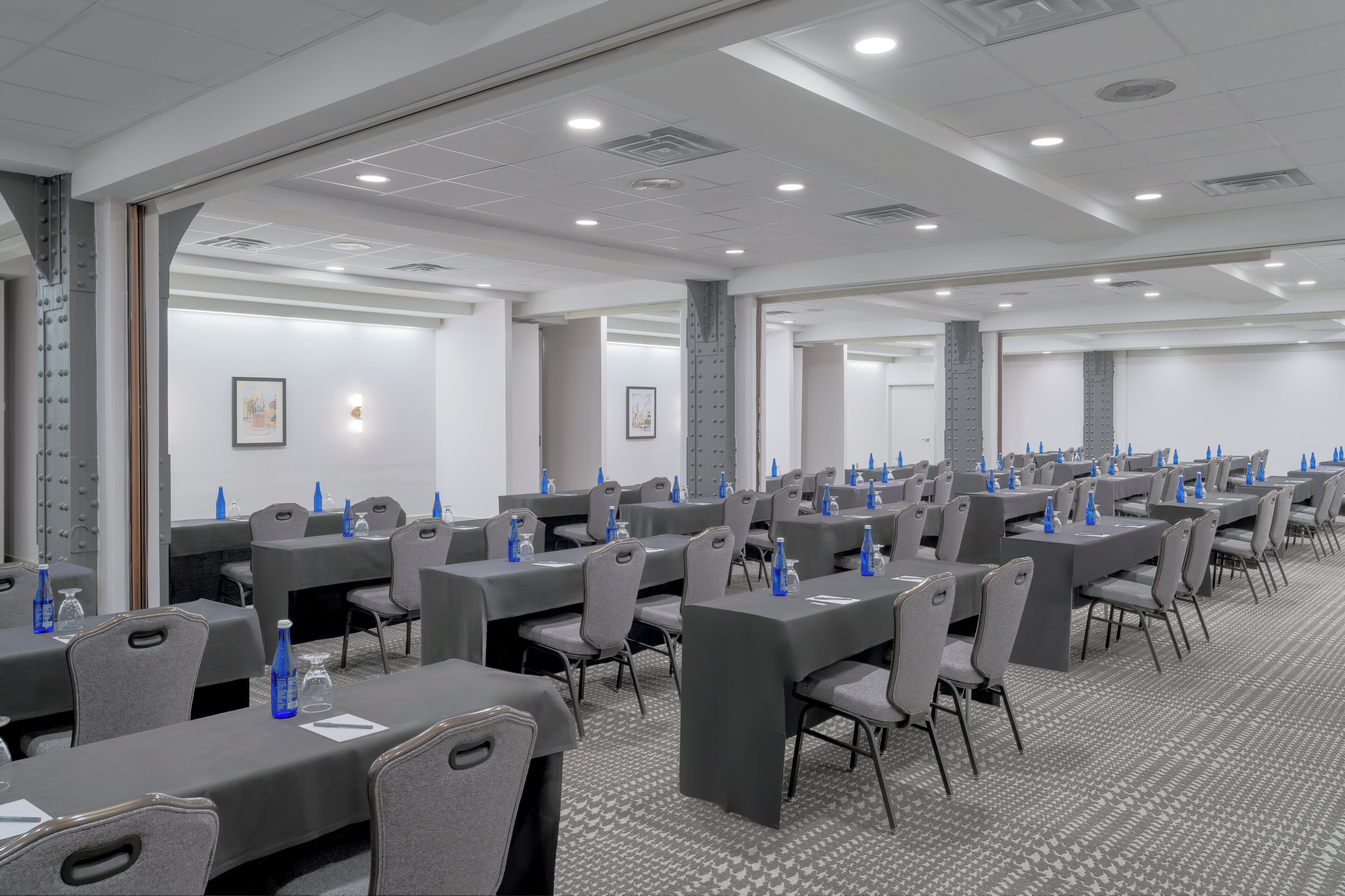 Un salón de conferencias con varias mesas y sillas, equipado con botellas de agua y vasos, y una alfombra estampada.