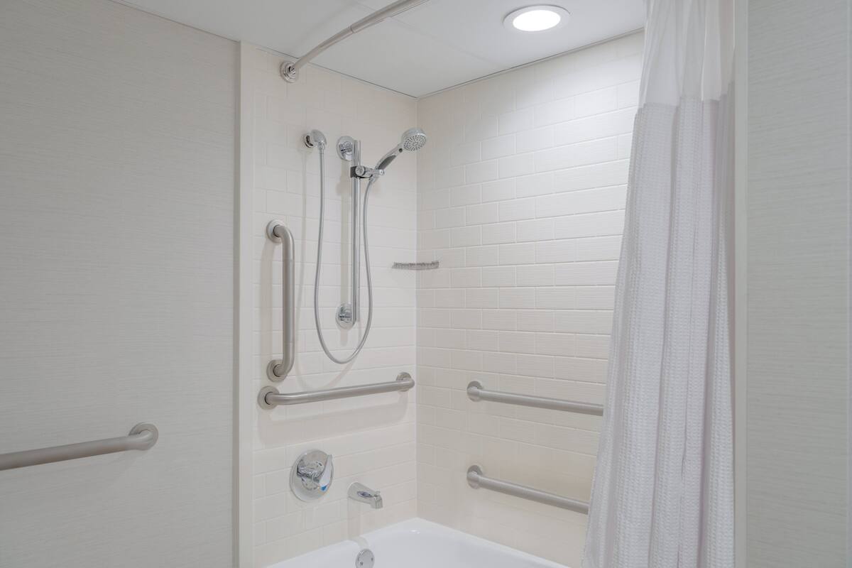 Un baño con paredes de azulejos blancos, una bañera blanca y una ducha con múltiples grifos y un cabezal de ducha de mano.