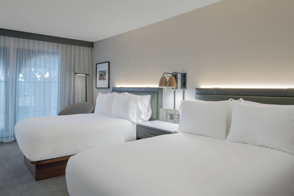 Habitación de hotel con dos camas, un escritorio, una silla, una lámpara y una ventana con cortinas.