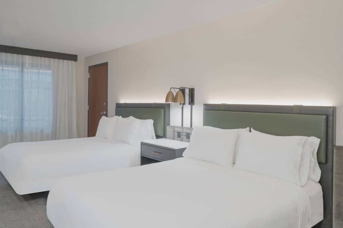Habitación de hotel con dos camas, una mesita de noche, una lámpara y un ventanal con cortinas.