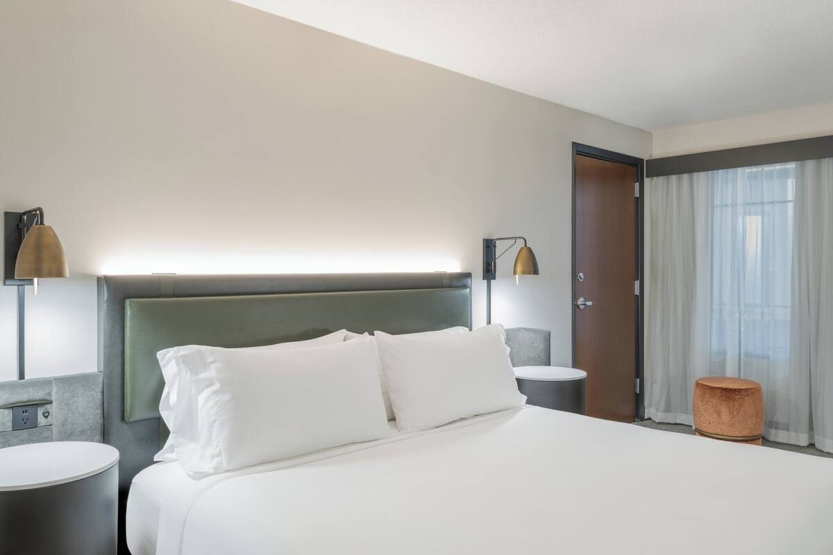 Una habitación de hotel con una cama grande, dos mesitas de noche, una lámpara, una puerta y una ventana con cortinas.