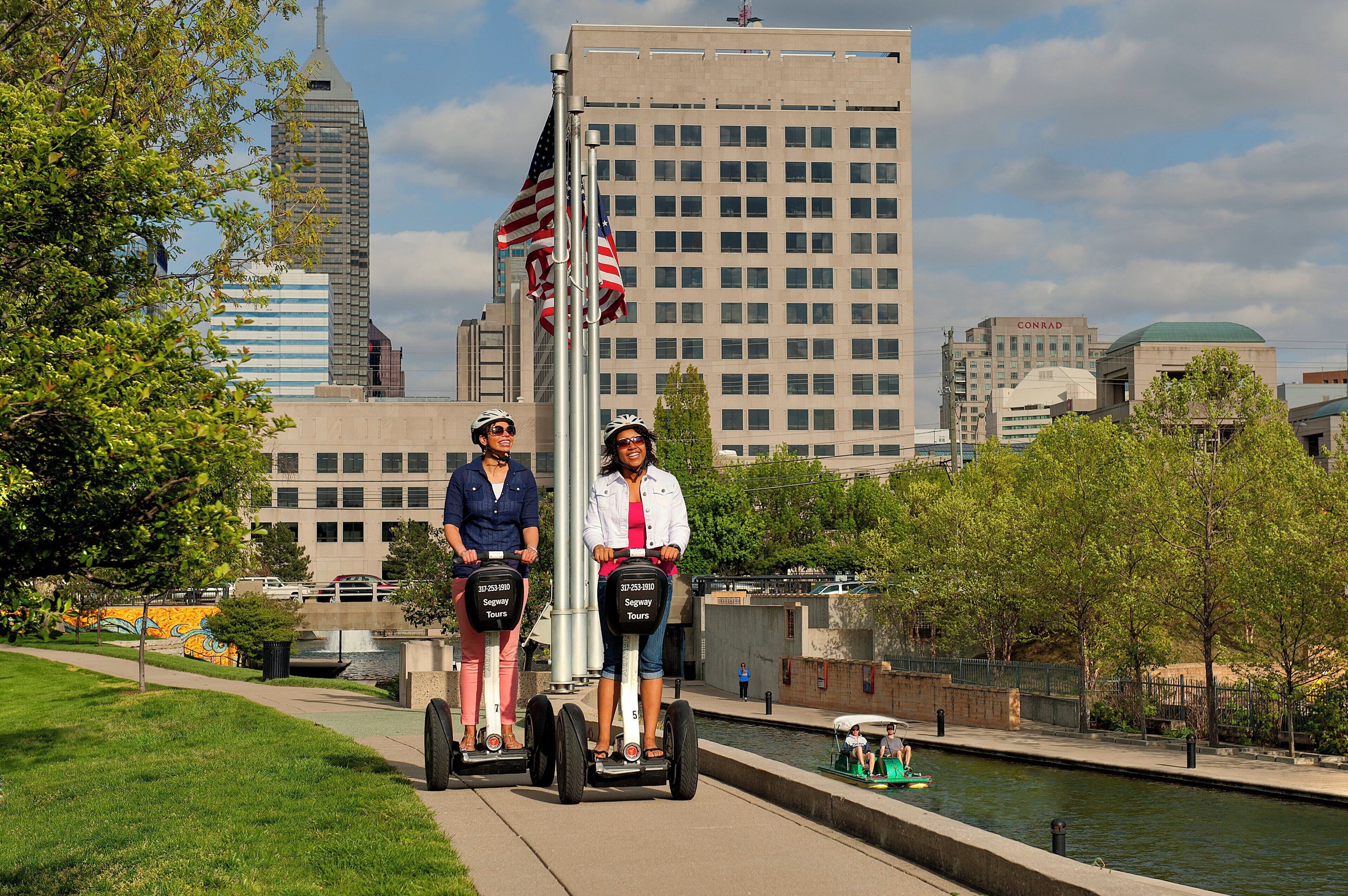 Dos personas realizan un recorrido en Segway en un entorno urbano, con edificios y un canal al fondo.