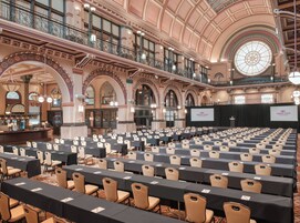 Un amplio salón de conferencias con filas de mesas y sillas, arquitectura ornamentada y un escenario con una pantalla que muestra el nombre del hotel.