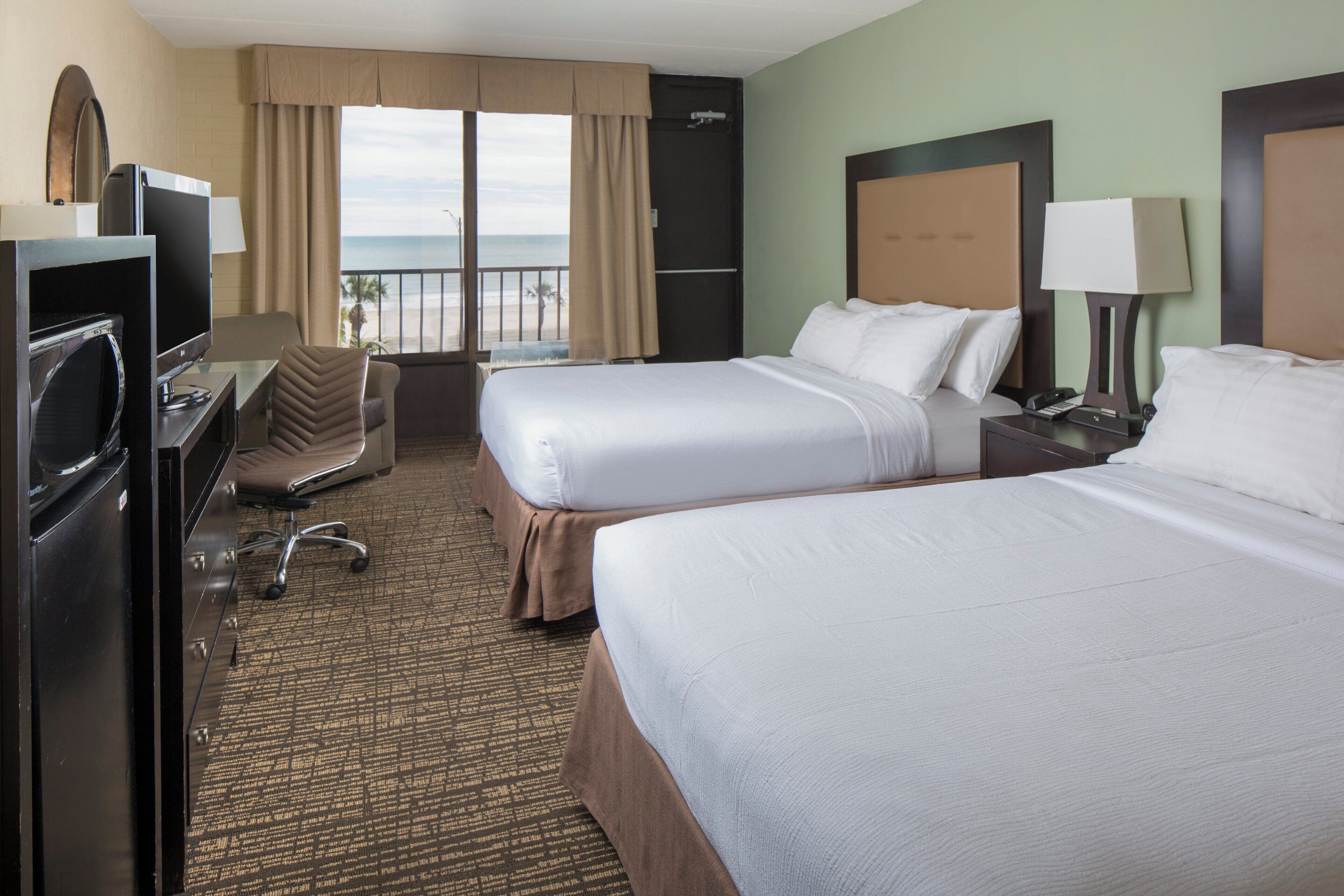 A hotel room with two beds, a desk, a chair, a television, and a view of the beach.