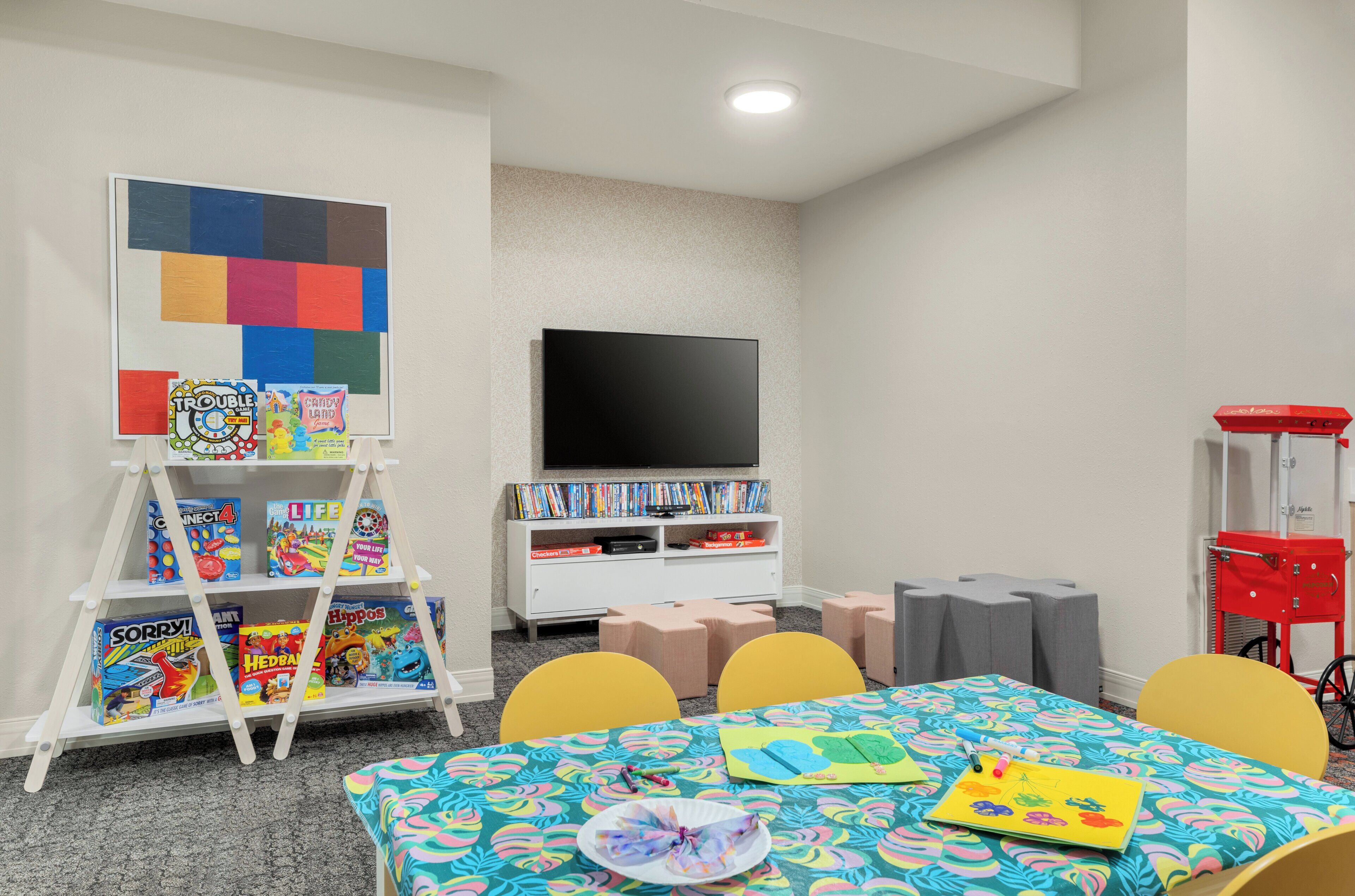 A children's playroom with a white bookshelf, colorful toys, a flat-screen TV, and a table with a patterned tablecloth.