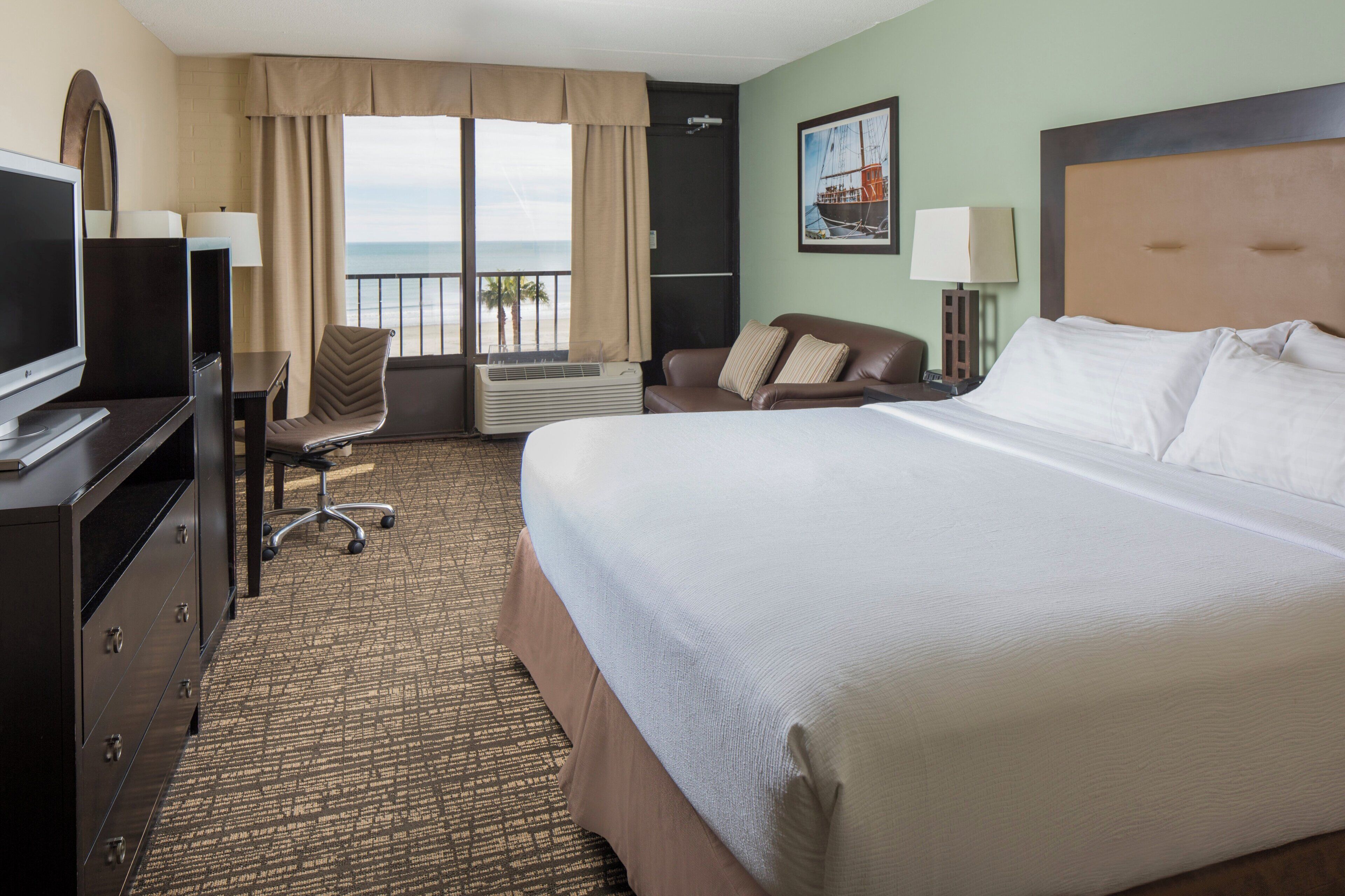 A hotel room with a large bed, a desk with a TV, a chair, a desk lamp, and a window with a view of the ocean.