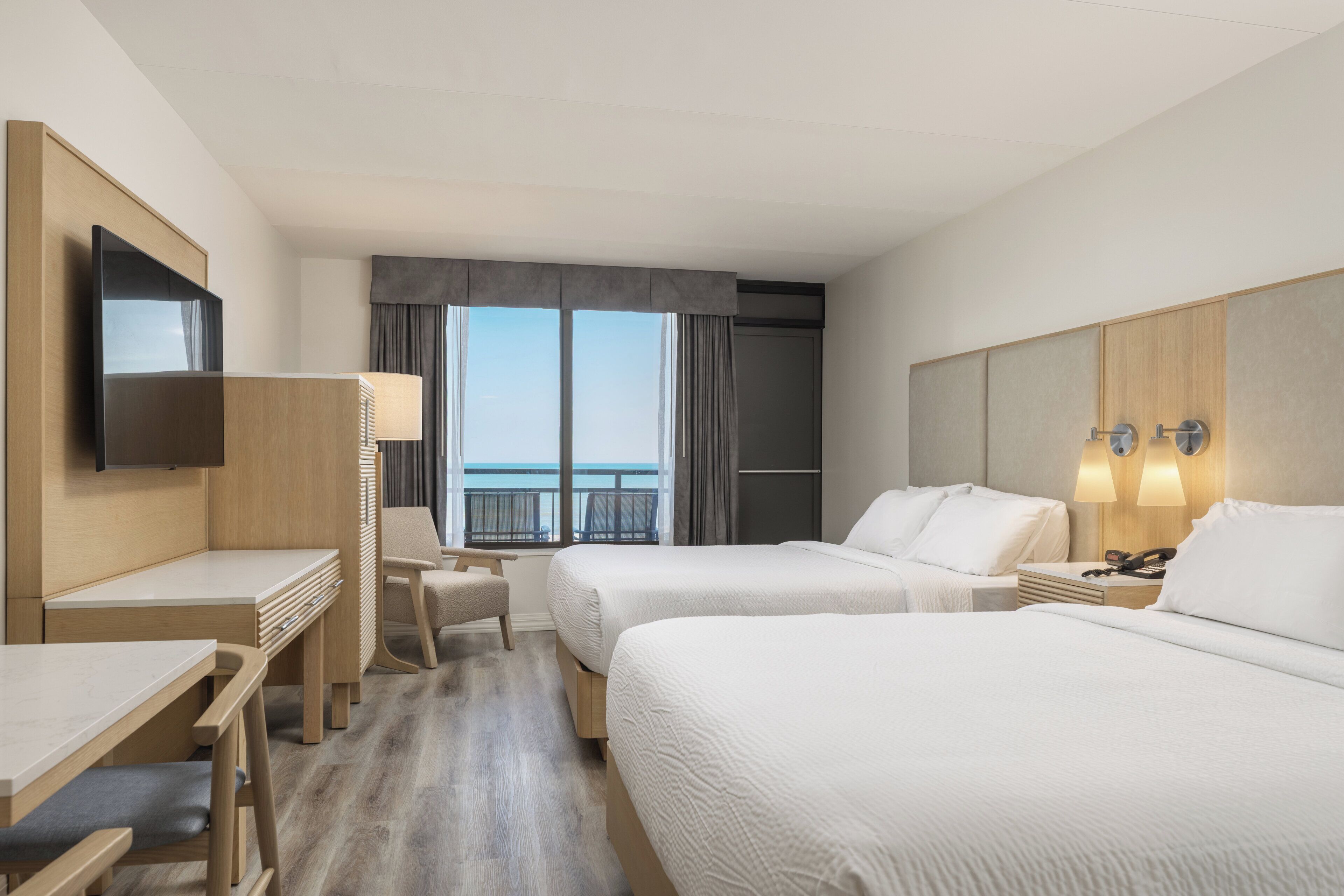 A hotel room with two beds, a desk, a chair, a TV, and a view of the ocean.