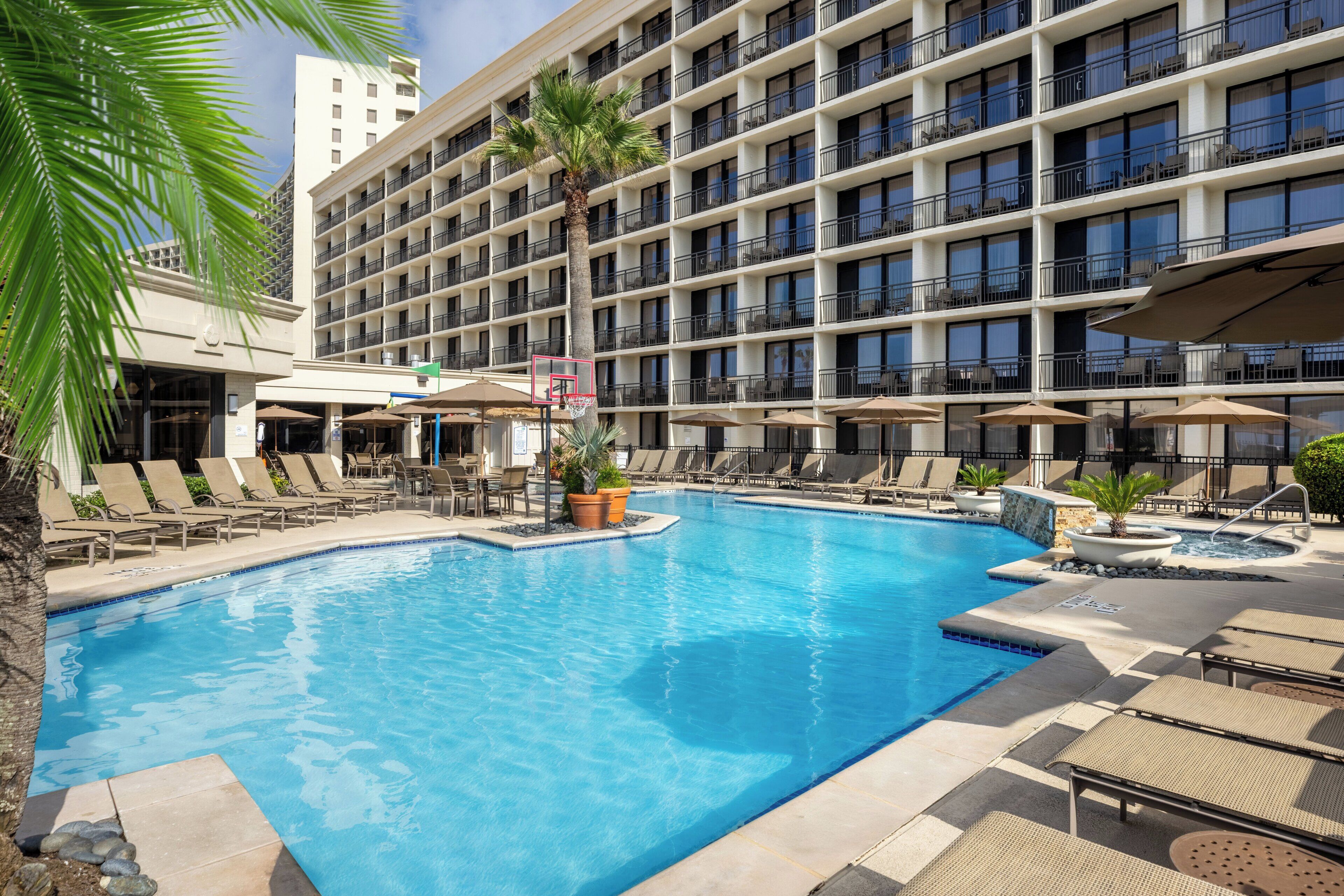 A hotel pool area with lounge chairs, umbrellas, and a basketball hoop.