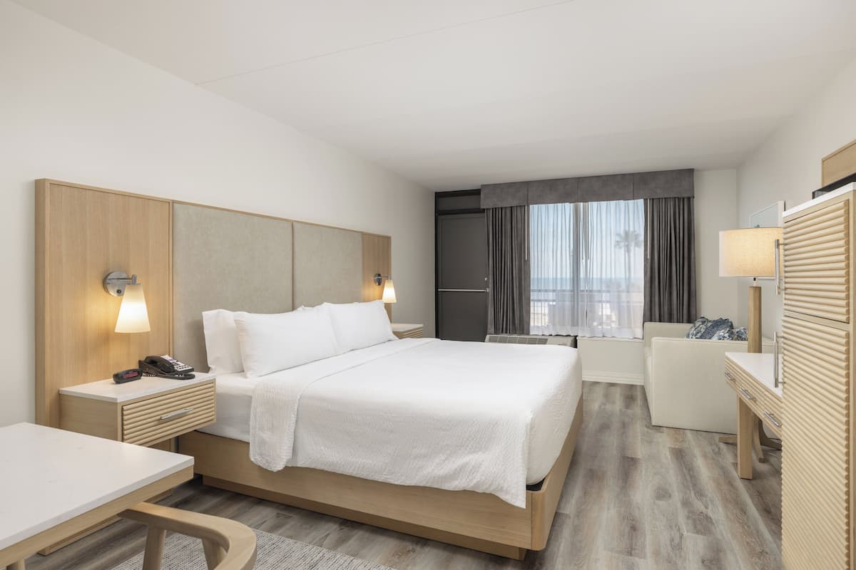 Ein modernes Hotelzimmer mit einem großen Bett, Nachttischen, einem Schreibtisch, einem Sessel und einer kleinen Küchenzeile, mit Blick auf die Stadt.