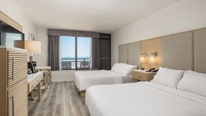 Quarto de hotel com duas camas, uma escrivaninha, uma cadeira, um abajur, um telefone e vista para o mar.