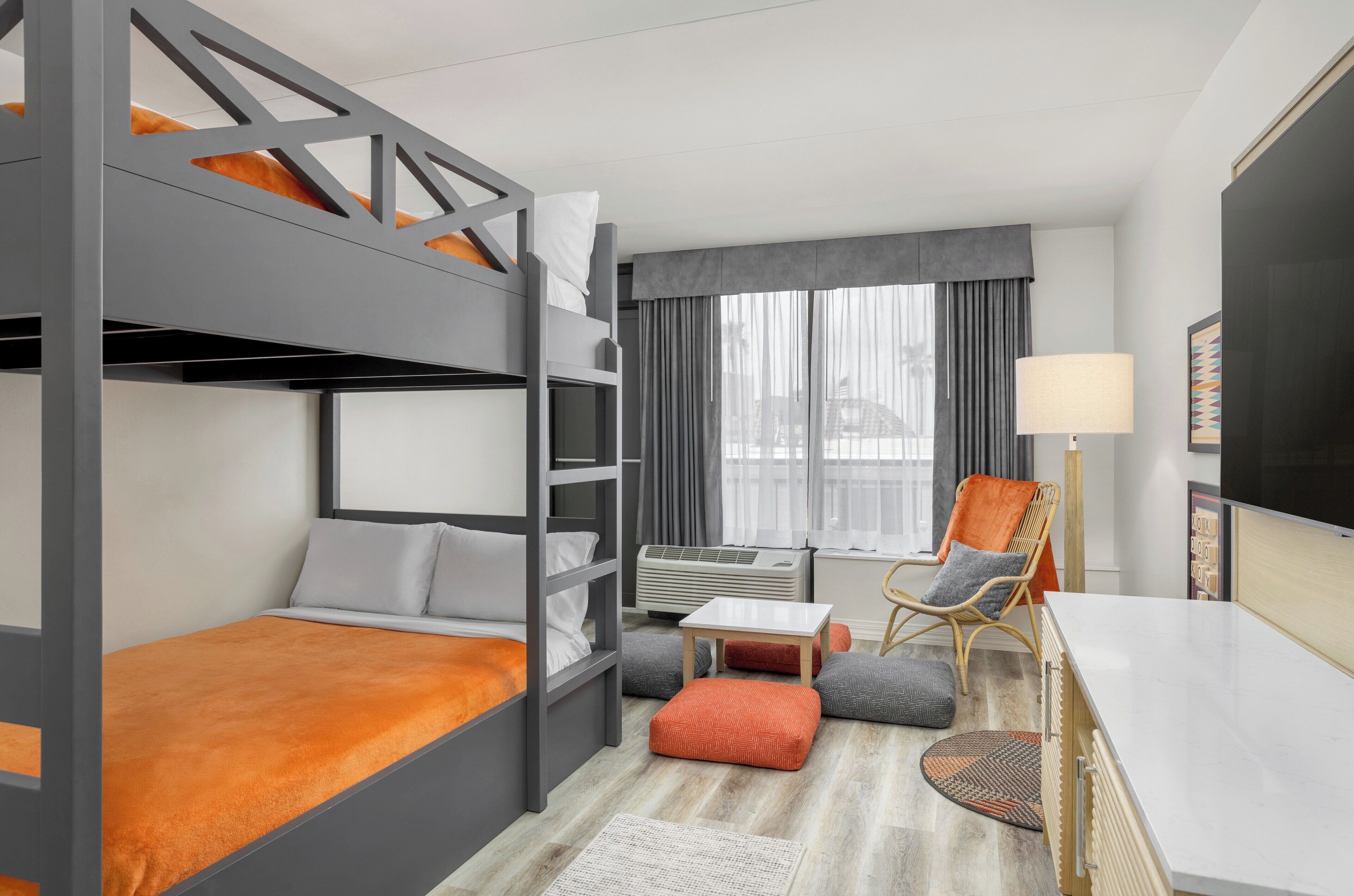 A modern hotel room with a bunk bed, a desk with a lamp, a chair, and a large window with curtains.