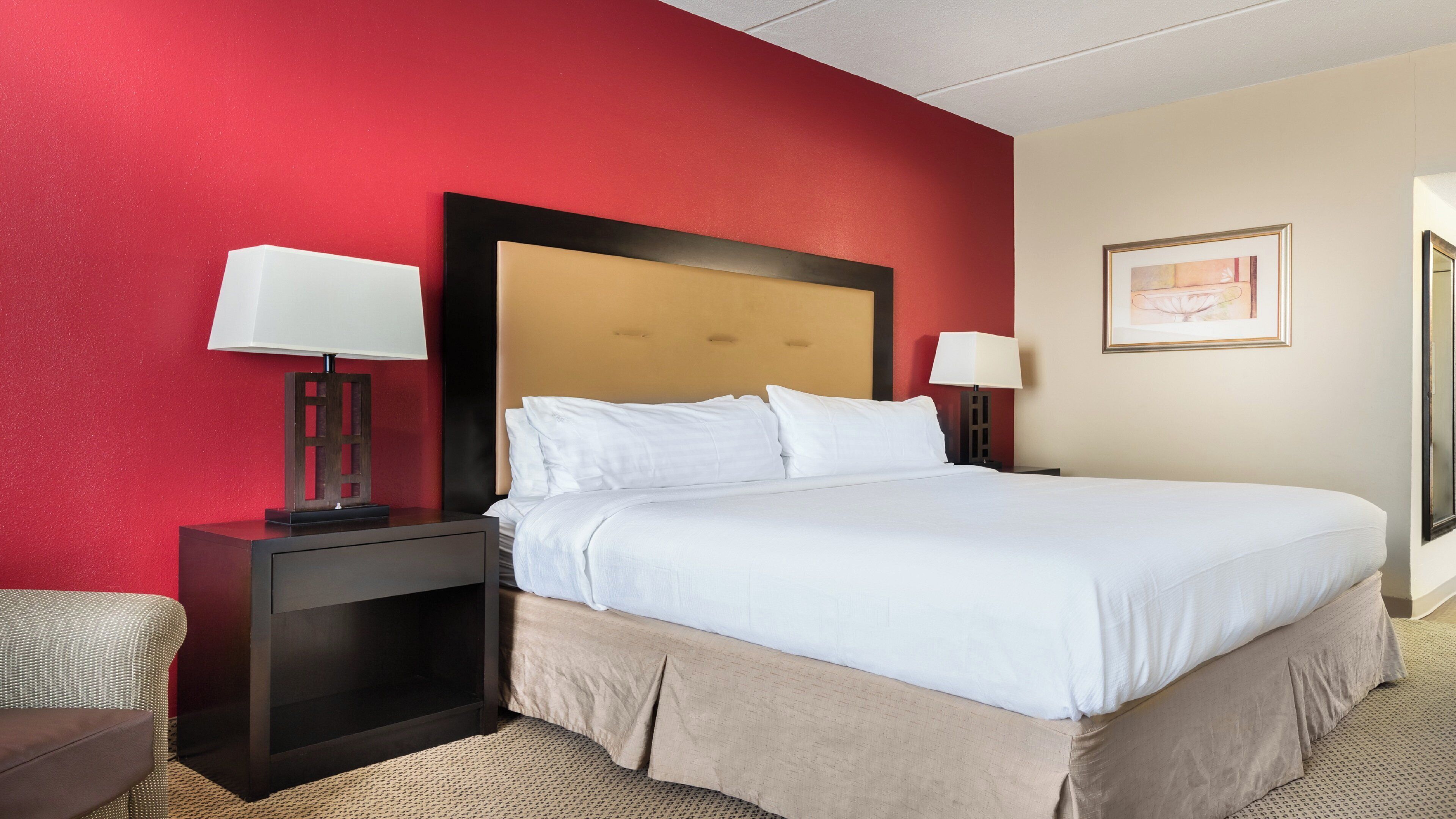 Ein Hotelzimmer mit einem großen Bett, zwei Nachttischen mit Lampen, einem Kopfteil, einem gerahmten Bild an der Wand und einer roten Akzentwand.