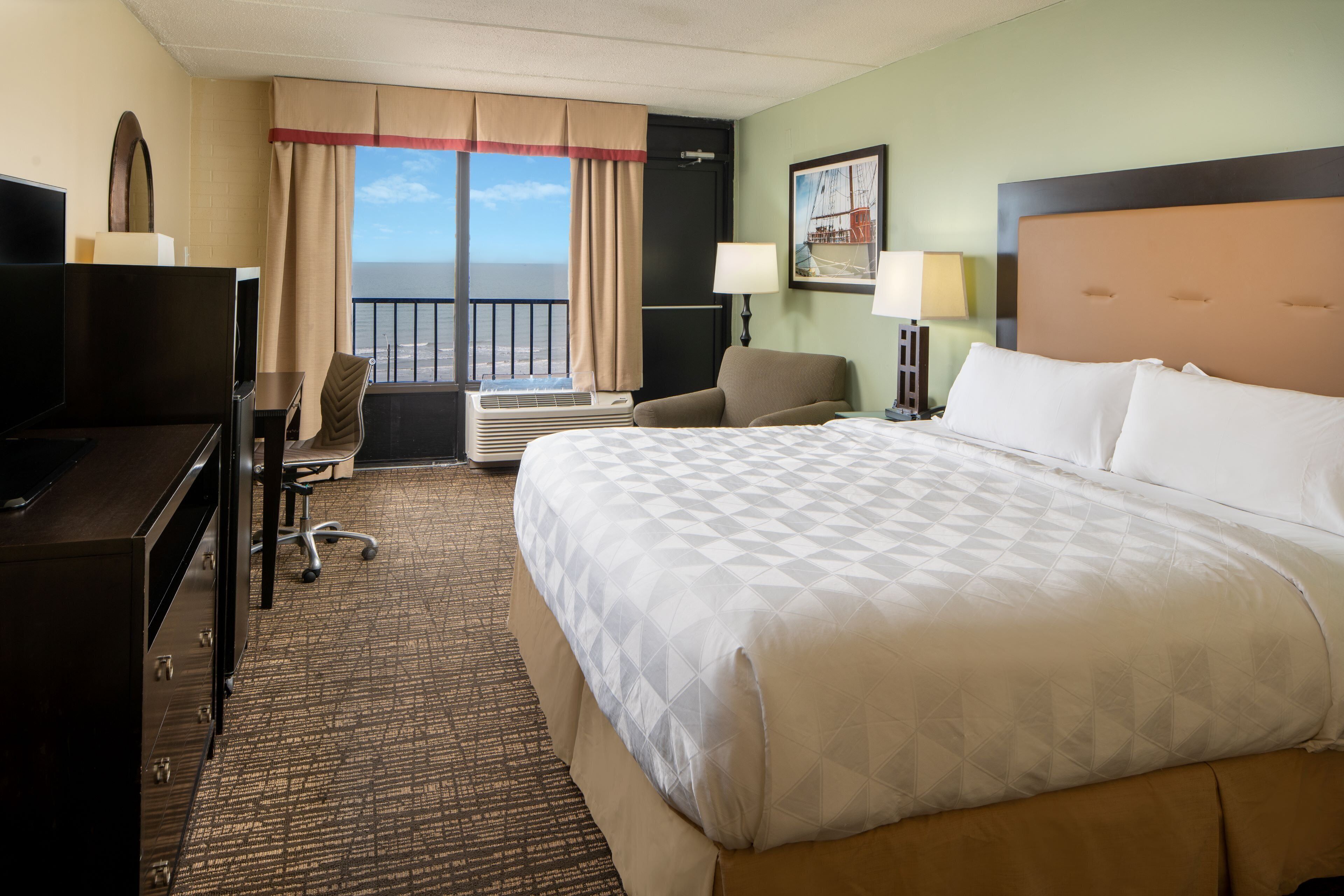 A hotel room with a large bed, a desk with a computer, a chair, an air conditioning unit, and a window with a view of the ocean.