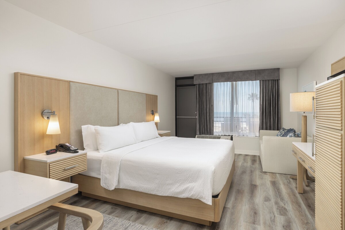Ein modernes Hotelzimmer mit einem großen Bett, Nachttischen, einem Schreibtisch, einem Sessel und einer kleinen Küchenzeile, mit Blick auf die Stadt.