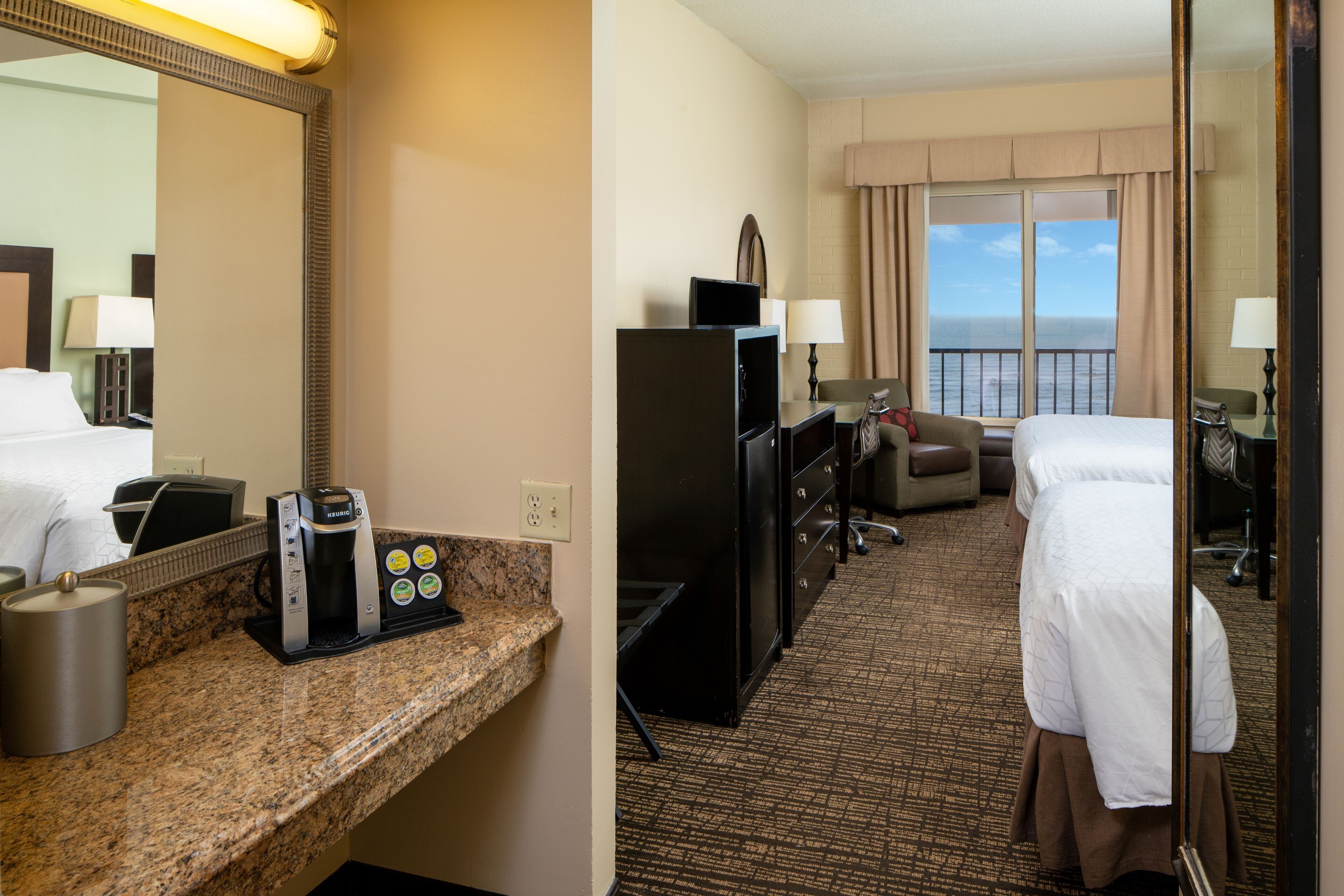 A hotel room with a bed, a desk with a coffee maker, a TV, and a view of the ocean.