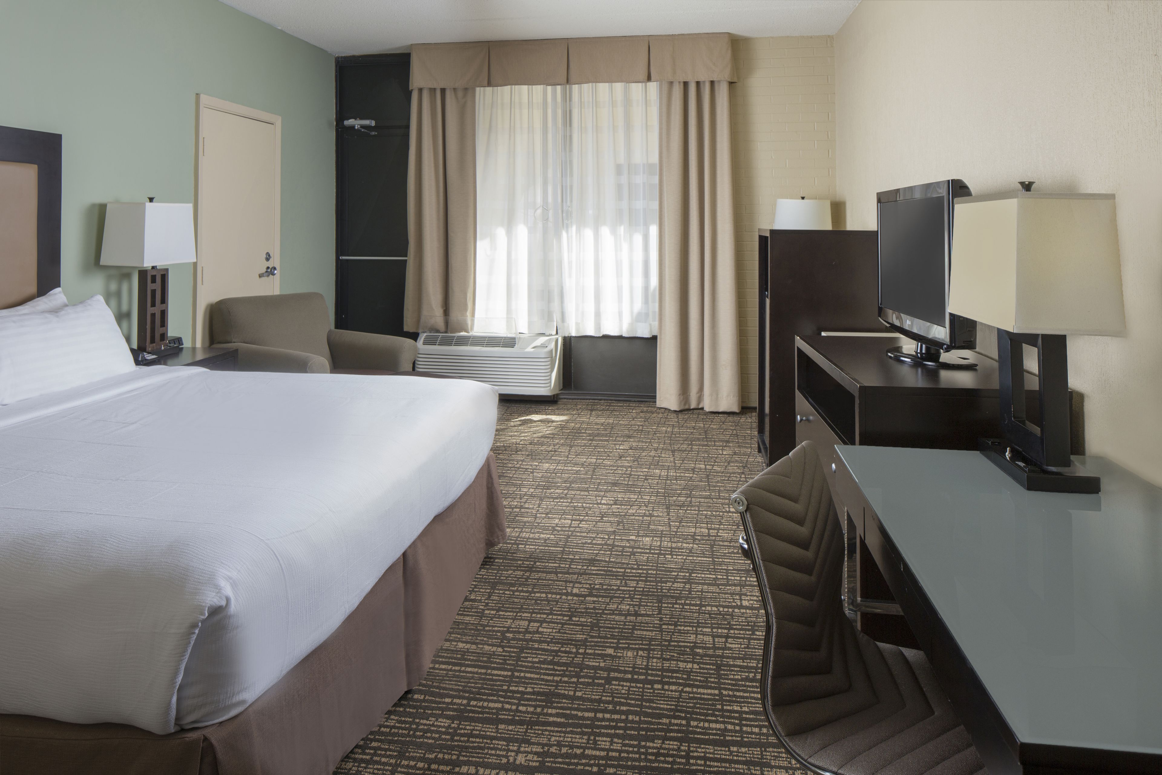 Une chambre d’hôtel avec un grand lit, un bureau avec une télévision, une chaise et une fenêtre avec des rideaux.