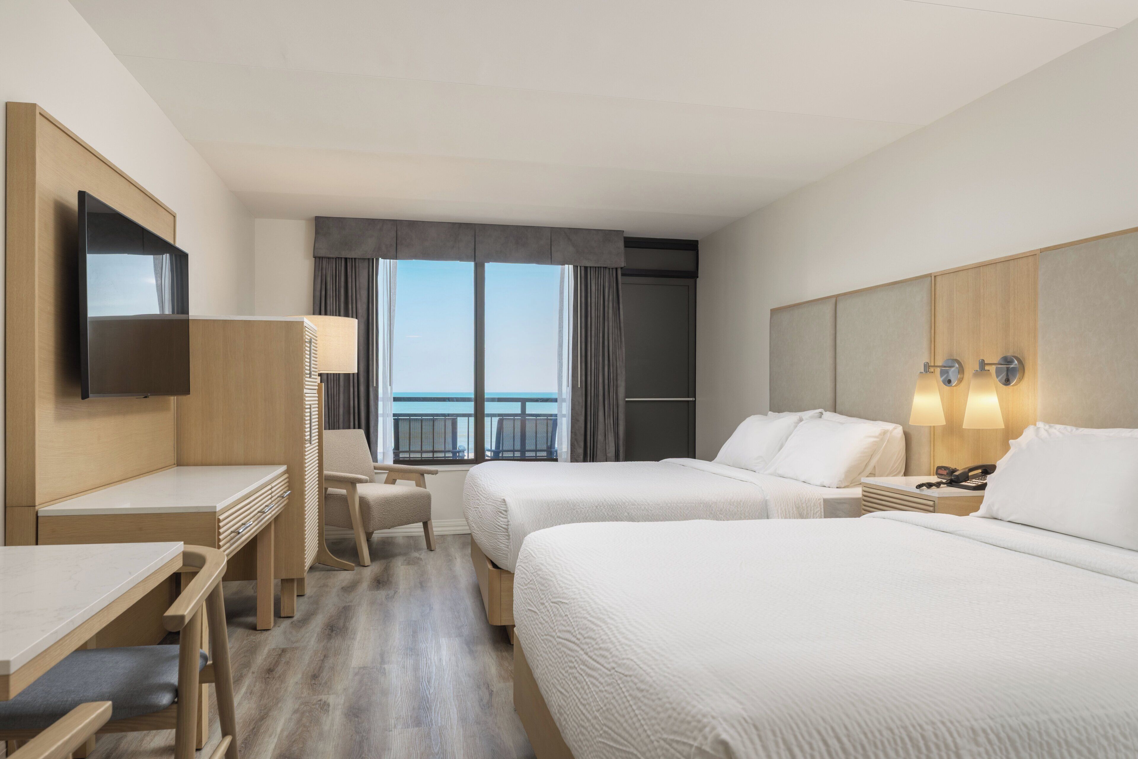 A hotel room with two beds, a desk, a chair, a TV, and a window with a view of the sea.