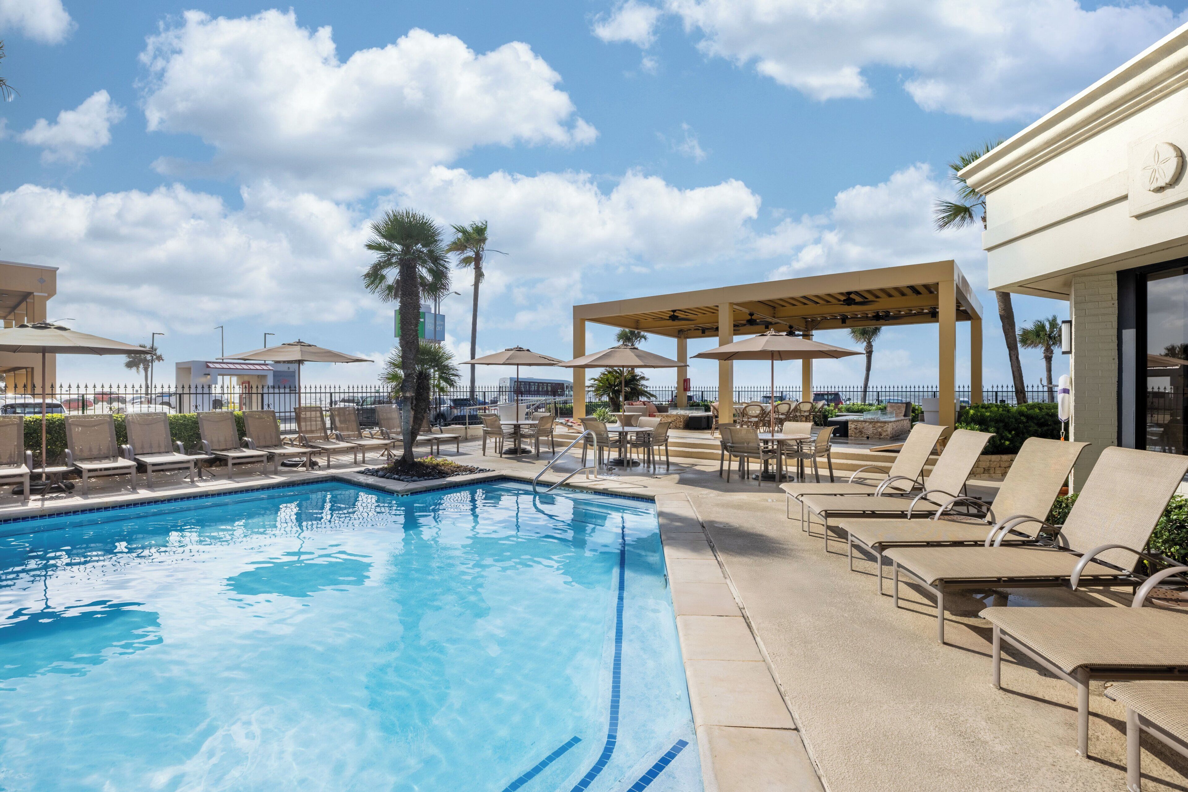 A pool area with lounge chairs and umbrellas, surrounded by a fence and palm trees.