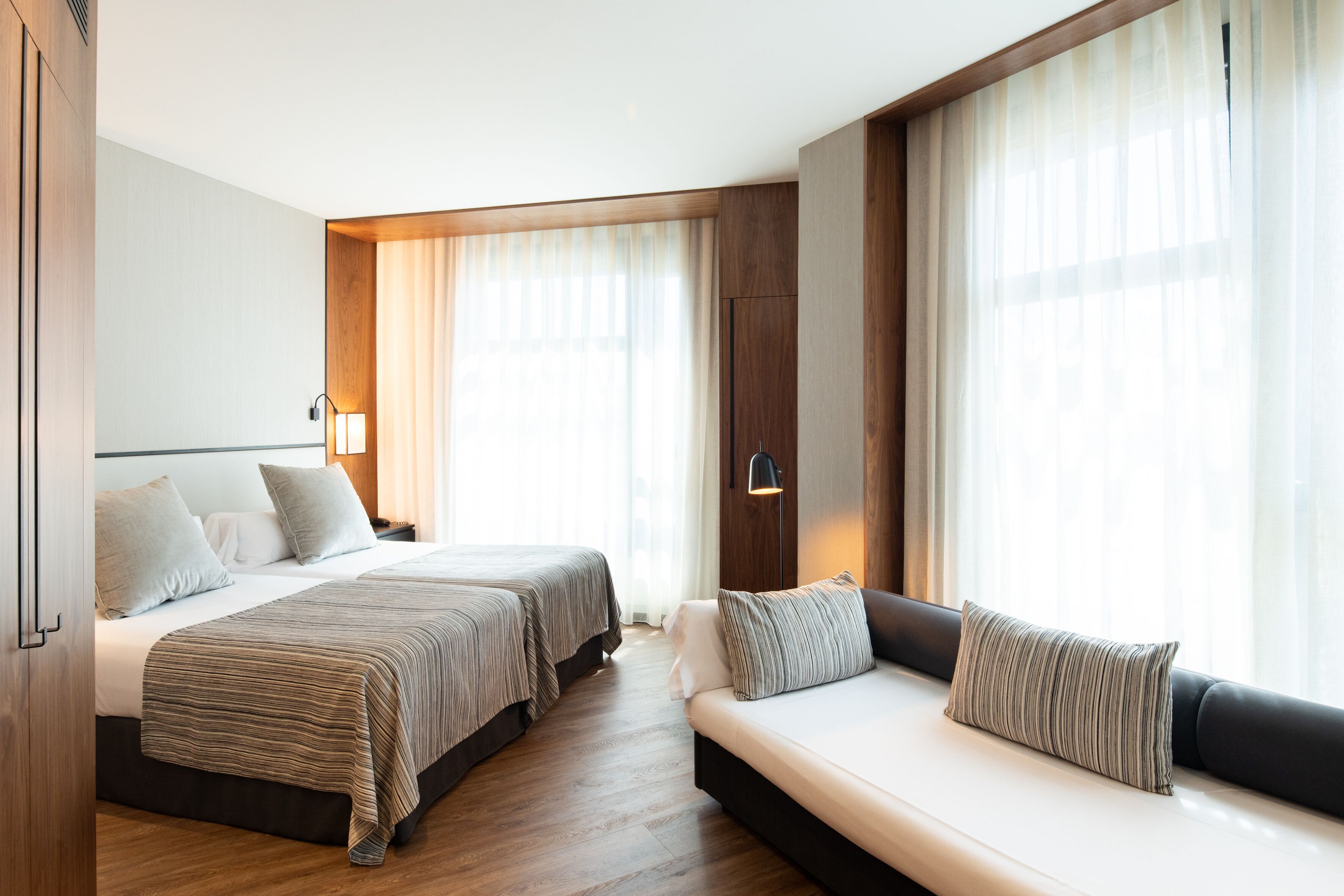Ein Hotelzimmer mit zwei Betten, einer Couch, Holzböden und großen Fenstern mit durchscheinenden Vorhängen.