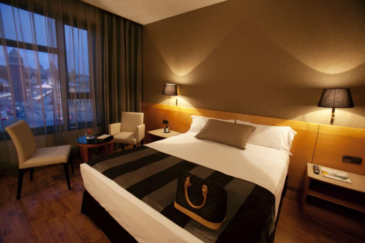 A hotel room with a large bed, two bedside tables, a chair, a small table, and a view of the cityscape through the window.