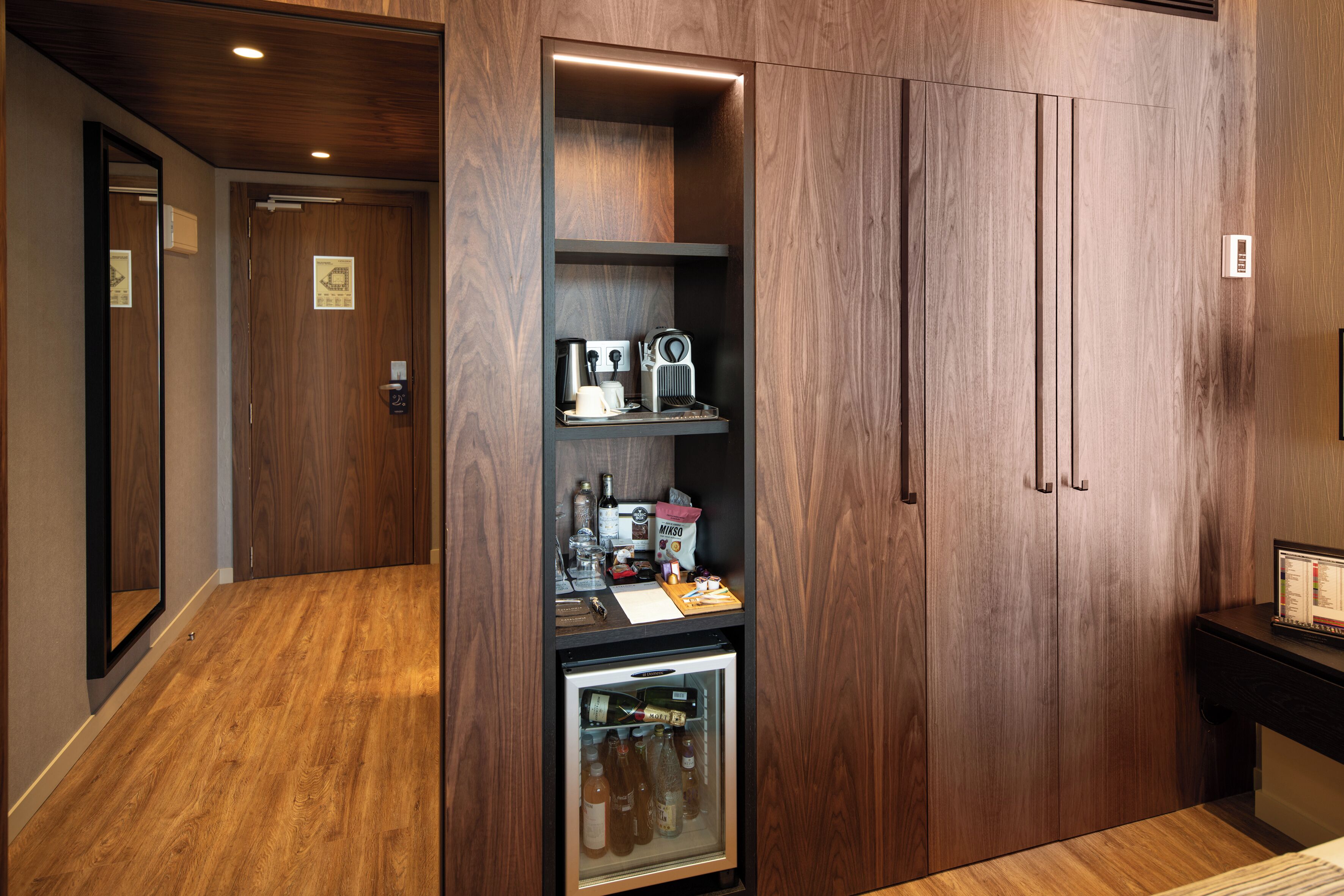 Una habitación de hotel con suelo de madera, una nevera con bebidas y aperitivos, una cafetera y un refrigerador.