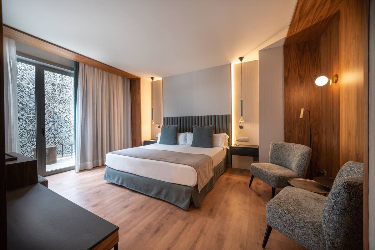 Une chambre d’hôtel moderne avec un grand lit, deux tables de chevet, un bureau et une chaise. On y trouve une grande fenêtre avec des rideaux, un panneau mural en bois et un luminaire au plafond.