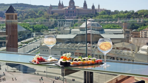 Une table en verre avec des sushis et deux verres d’eau, donnant sur un paysage urbain avec des bâtiments historiques.