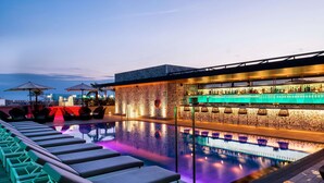 Un espace piscine sur le toit, comprenant des chaises longues, un bar et une piscine illuminée de couleurs vives.