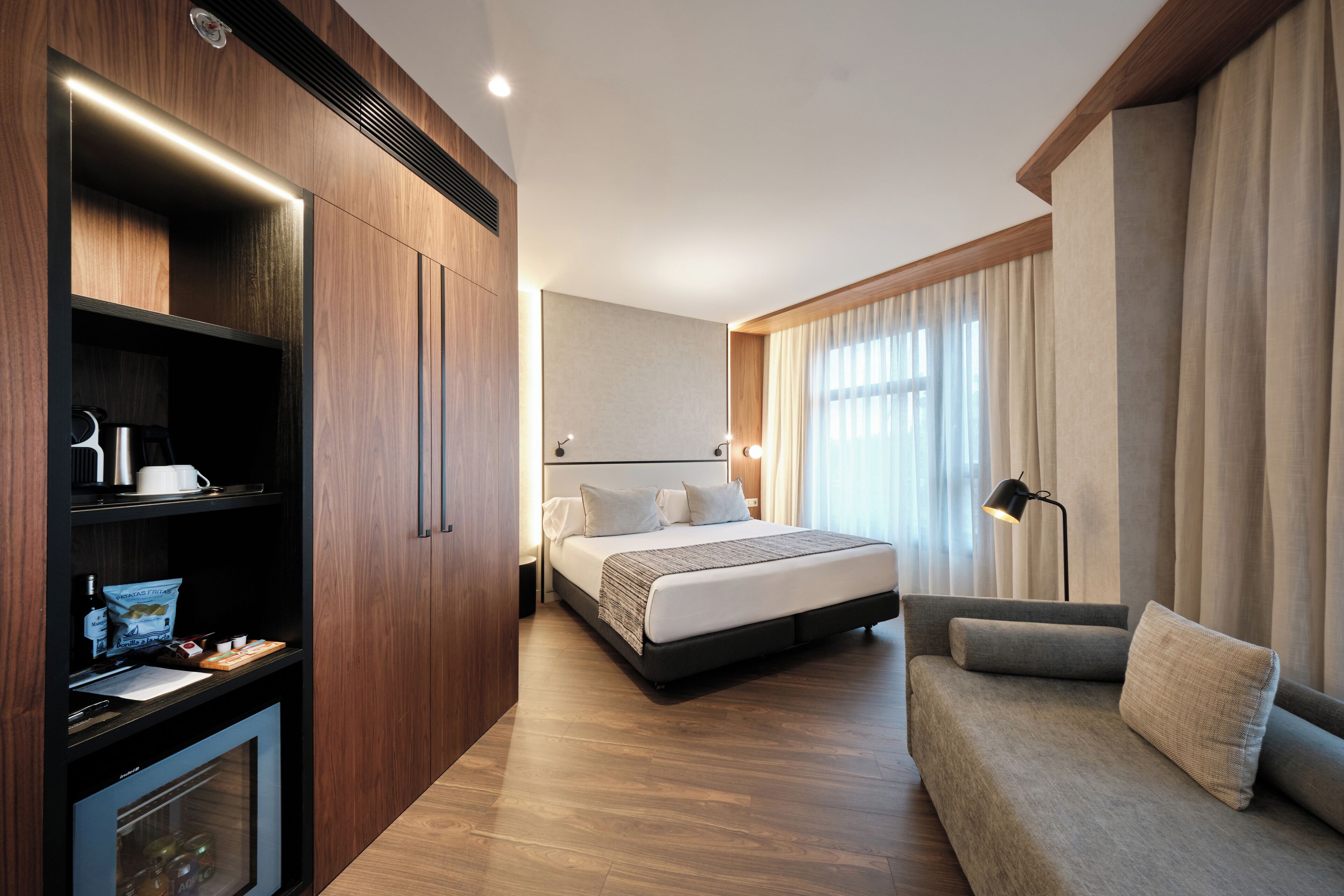 A modern hotel room with a wooden wardrobe, a bed with a grey upholstered sofa, a desk with a lamp, and a large window with curtains.
