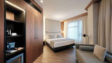 A modern hotel room with a wooden wardrobe, a bed with a grey upholstered sofa, a desk with a lamp, and a large window with curtains.