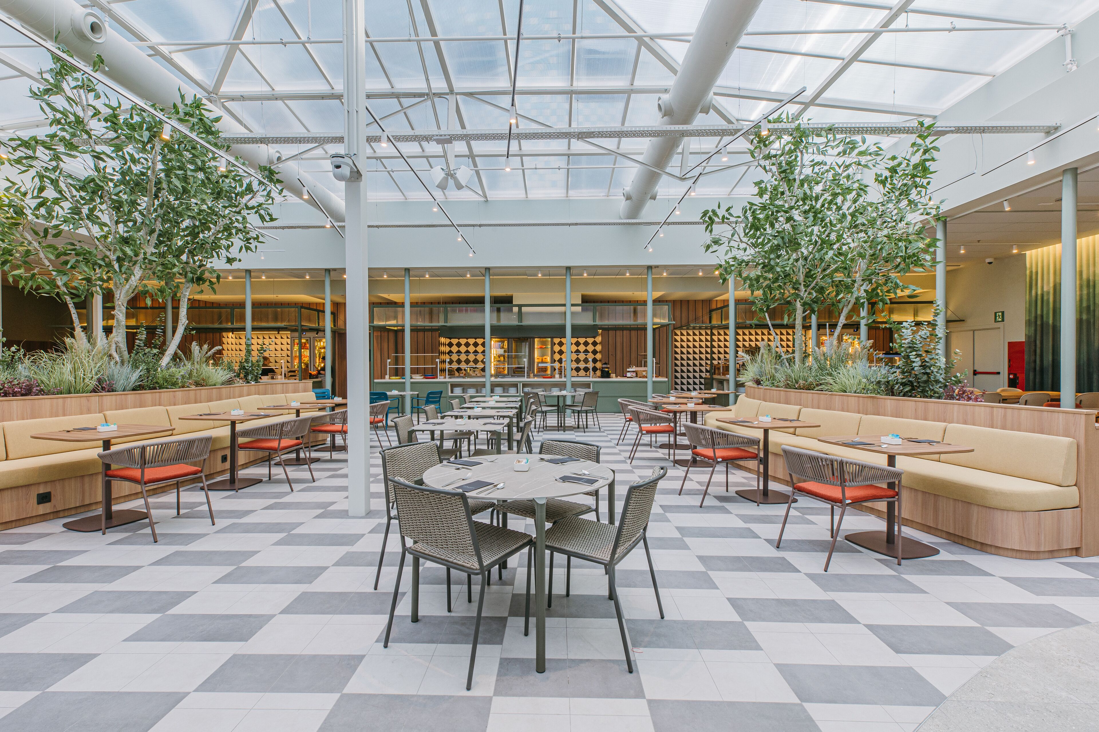 A spacious indoor area with multiple seating arrangements, including tables and sofas, surrounded by potted plants and a high ceiling with natural light.